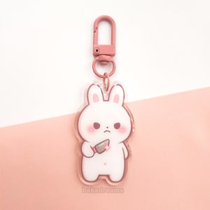 May include: A clear acrylic keychain featuring a white cartoon bunny holding a knife. The bunny has pink cheeks and a stern expression. The keychain has a pink clasp and ring. The text "bakadreams" is at the bottom.