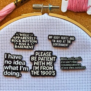 May include: A cross-stitch project in a wooden hoop, with several black and white text patches pinned to the fabric. The patches feature humorous phrases such as "I have no idea what I'm doing" and "Please be patient with me, I'm from the 1900's."