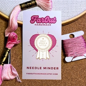 May include: A pink and white needle minder with the text "Only Cried A Little" and "Yay" on a white card. The card is next to pink embroidery floss and a wooden embroidery hoop.
