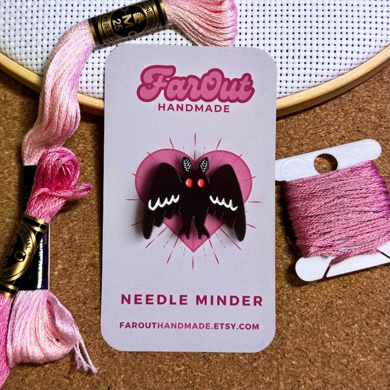 May include: Black needle minder shaped like a bat with red eyes. The needle minder is on a white card with the text "FarOut Handmade" and "Needle Minder" in pink and black lettering. The card also includes the website "farouthandmade.etsy.com".
