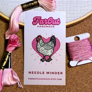Peut inclure: Un protège-aiguille rose et blanc avec une illustration de poule dans un cœur rose. La carte indique "Far Out Handmade" et "Needle Minder" avec une adresse de site web. Du fil à broder rose et un cercle à broder en bois sont en arrière-plan.