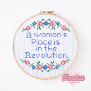 May include: A white cross-stitch embroidery hoop with a blue and pink floral border. The text "A woman's place is in the revolution." is stitched in blue thread.