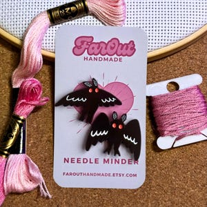 May include: Two black needle minders shaped like moths with red eyes and white markings. The minders are on a white card with the text "FarOut Handmade" and "Needle Minder" in black. The card also has a pink heart shape and the website address "farouthandmade.etsy.com".