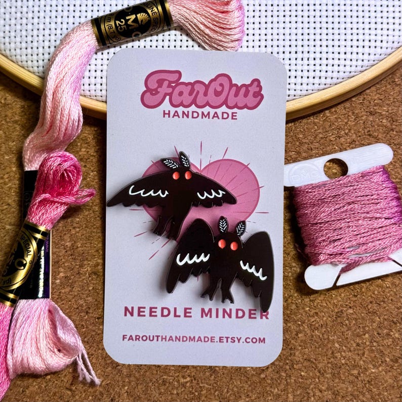 May include: Two black needle minders shaped like moths with red eyes and white markings. The minders are on a white card with the text "FarOut Handmade" and "Needle Minder" in black. The card also has a pink heart shape and the website address "farouthandmade.etsy.com".