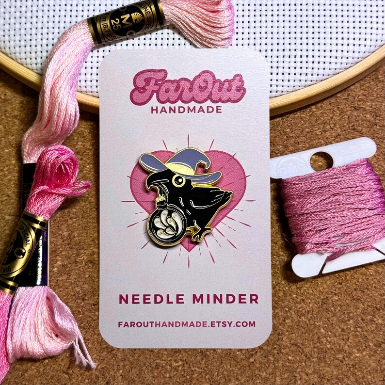 K&ouml;nnte beinhalten: Eine schwarze Kr&auml;he mit einem lila Hexenhut, die einen goldenen Nadelhalter mit wei&szlig;en Federn h&auml;lt. Die Kr&auml;he befindet sich auf einem rosafarbenen Herzhintergrund mit dem Text "FarOut Handmade" und "Needle Minder" auf einer wei&szlig;en Karte.