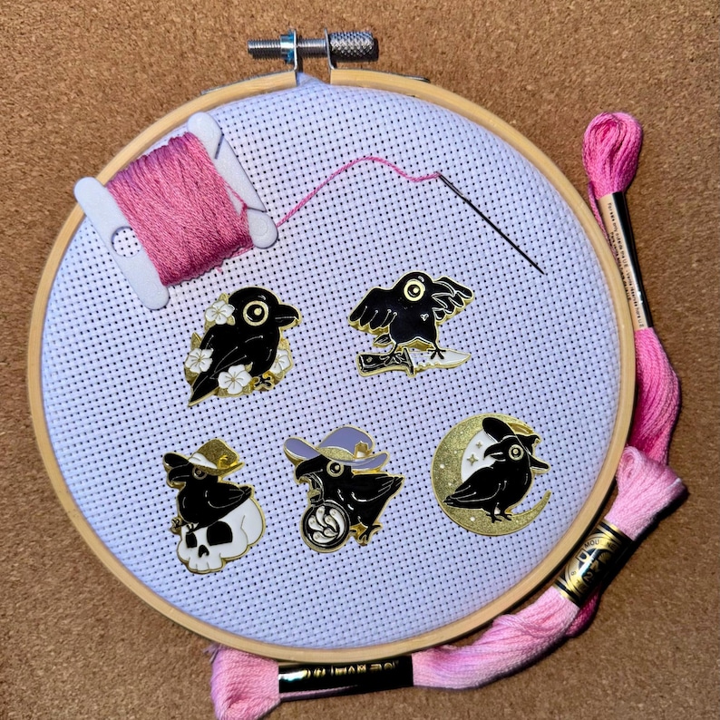 K&ouml;nnte beinhalten: Ein wei&szlig;er Stickrahmen mit rosa Stickgarn und sechs schwarzen Raben-Pins. Die Pins zeigen verschiedene Designs, darunter einen Raben mit einem Sch&auml;del, einen Raben mit einem Mond und einen Raben mit einem Hexenhut.