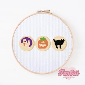 May include: A white embroidery hoop with a beige background and three cross-stitch designs. The designs are a purple ghost, an orange pumpkin, and a black cat.