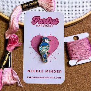 Peut inclure: Épingle à nourrice représentant un geai bleu, bordée d'or, sur un cœur rose et une carte blanche avec le texte "Far Out Handmade" et "Needle Minder". Du fil à broder rose et un cercle à broder en bois en arrière-plan.