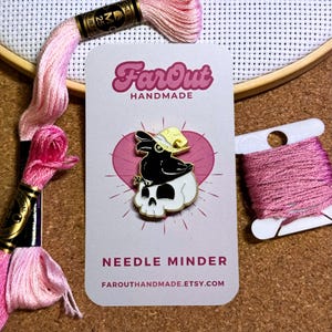 K&ouml;nnte beinhalten: Eine schwarze und wei&szlig;e Emaille-Nadel mit einer Kr&auml;he, die einen gelben Hut tr&auml;gt und auf einem Sch&auml;del sitzt. Die Nadel befindet sich auf einer wei&szlig;en Karte mit dem Text "FarOut Handmade" und "Needle Minder" sowie der Website "farouthandmade.etsy.com".