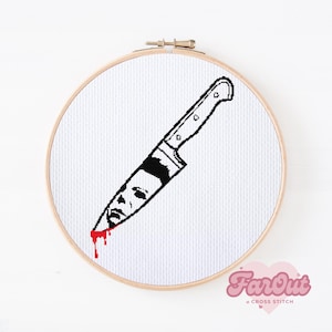 May include: A white embroidery hoop with a black and white cross stitch design of a knife with a face on the blade and a red drip of blood. The design is on a white fabric background.
