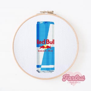 Può includere: Un punto croce di una lattina di Red Bull in un telaio da ricamo in legno. La lattina è blu e bianca con il logo Red Bull in rosso. Le parole "SUGAR FREE" sono sotto. Lo sfondo è bianco.