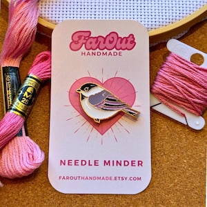 Peut inclure: Épingle à broder en forme d'oiseau rose et doré, avec le texte "Far Out Handmade" et "Needle Minder". L'épingle est posée sur un cœur rose. Du fil à broder rose et un cercle à broder en bois sont visibles en arrière-plan.