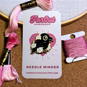 K&ouml;nnte beinhalten: Schwarze Kr&auml;he Nadelhalter mit wei&szlig;en Blumen, goldenen Akzenten und dem Text "FarOut Handmade" und "Needle Minder" auf einer wei&szlig;en Karte mit einem rosa Herzen.