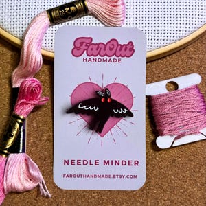 May include: A black needle minder shaped like a moth with red eyes. The needle minder is on a white card with a pink heart and the text "FarOut Handmade" and "Needle Minder" in black. The text "FarOutHandmade.etsy.com" is also on the card.