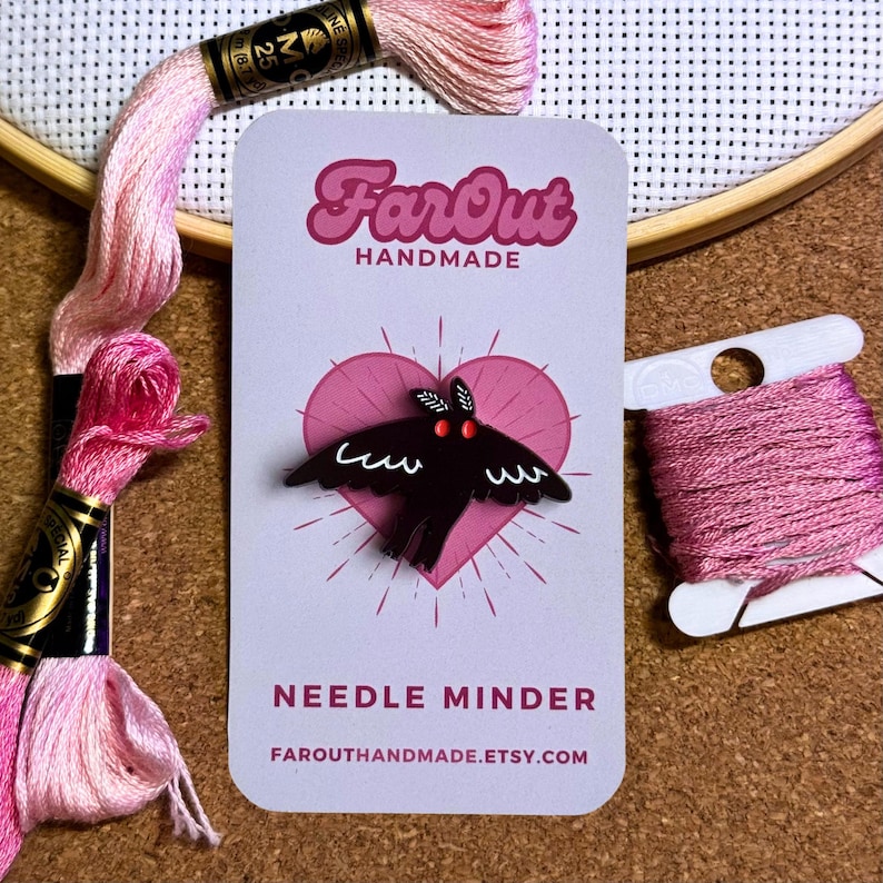 May include: A black needle minder shaped like a moth with red eyes. The needle minder is on a white card with a pink heart and the text "FarOut Handmade" and "Needle Minder" in black. The text "FarOutHandmade.etsy.com" is also on the card.