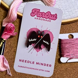 May include: A black bat needle minder with red eyes and a needle threaded through it. The minder is on a pink heart-shaped background with the text "FarOut Handmade" and "Needle Minder" on the card. The text "FarOutHandmade.etsy.com" is also on the card.