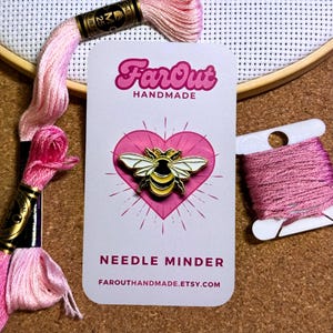 May include: A gold bee needle minder with white wings and a yellow and black striped body. The bee is on a white card with a pink heart and the text "FarOut Handmade" and "Needle Minder" with the website "farouthandmade.etsy.com".