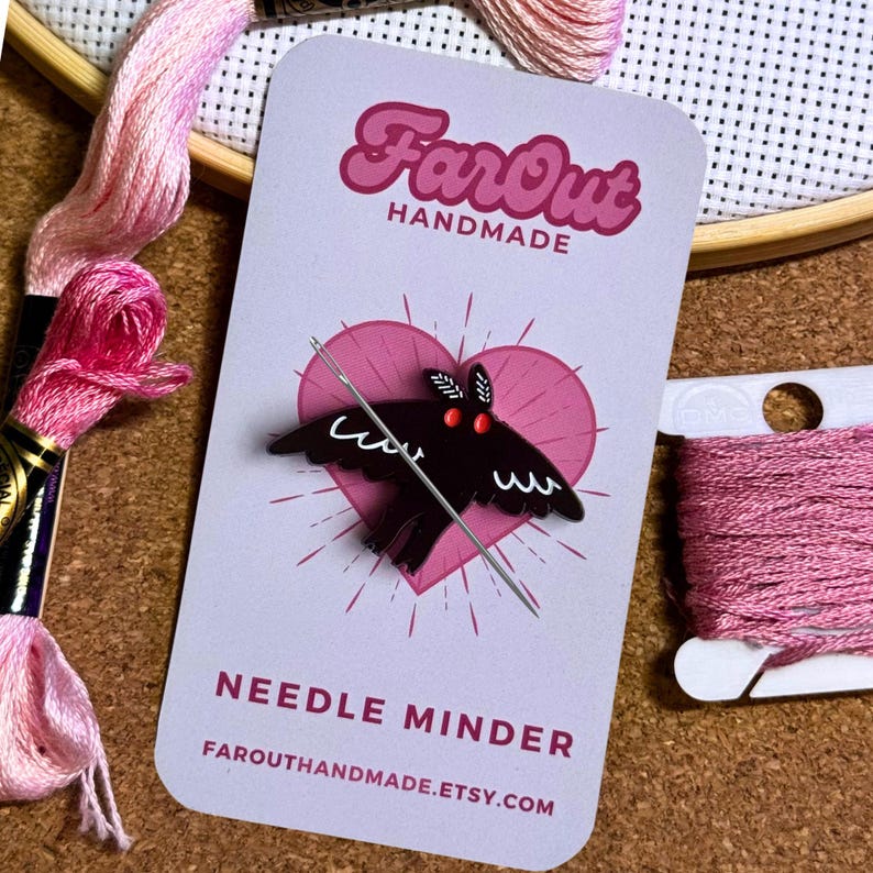 May include: A black bat needle minder with red eyes, a needle threaded through it, and the text "FarOut Handmade" and "Needle Minder" on a pink and white card.