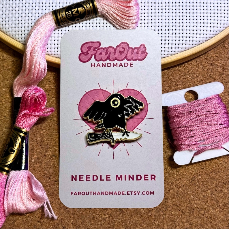 K&ouml;nnte beinhalten: Schwarze Emaille-Nadel mit einem Raben, der auf einem Messer steht, mit den Worten "Needle Minder" und "FarOutHandmade.etsy.com" auf einer wei&szlig;en Karte mit einem rosa Herzen.