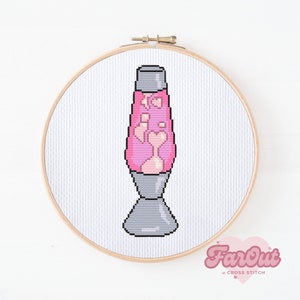 Op de afbeelding: Een roze lava lamp met een grijze voet, gekruist gestikt op witte stof in een houten borduurring. De stof is gelabeld "FarOut Cross Stitch".