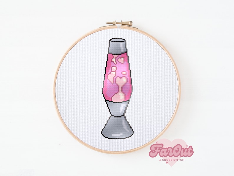 Op de afbeelding: Een roze lava lamp met een grijze voet, gekruist gestikt op witte stof in een houten borduurring. De stof is gelabeld "FarOut Cross Stitch".