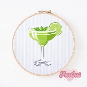 May include: A cross stitch pattern of a green margarita cocktail in a glass with a lime wedge garnish. The pattern is in a wooden embroidery hoop.