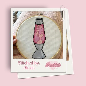 Op de afbeelding: Een kruissteek van een roze en witte lava lamp in een houten borduurring. De tekst "Stitched by: Alexis FarOut Cross Stitch" staat onder de ring.