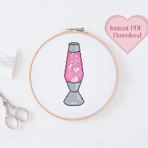Op de afbeelding: Een roze en witte lava lamp kruissteekpatroon in een houten borduurring. Het patroon is in een gepixelde stijl. De tekst "Direct PDF downloaden" staat in een roze hart.