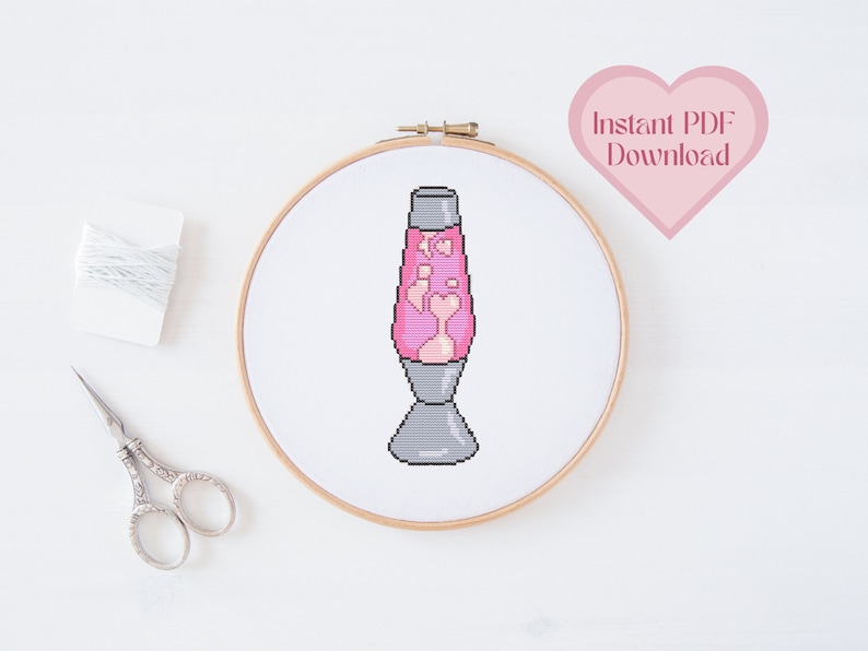 Op de afbeelding: Een roze en witte lava lamp kruissteekpatroon in een houten borduurring. Het patroon is in een gepixelde stijl. De tekst "Direct PDF downloaden" staat in een roze hart.