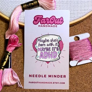 May include: A pink and purple needle minder with the text "Maybe she's born with it, MAYBE IT'S ADHD". The needle minder is on a white card with the text "FarOut Handmade" and "Needle Minder". Pink embroidery floss and a wooden hoop are in the background.
