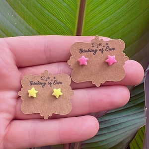 May include: Two pairs of star-shaped stud earrings displayed on brown scalloped cardboard cards. One pair is bright yellow, and the other pair is vibrant pink. Each card features the printed text Thinking of Ewe with a small sheep icon.