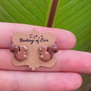 May include: A pair of brown squirrel-shaped stud earrings attached to a scalloped tan cardboard display card. The card features the text Thinking of Ewe with a small sheep icon and stars printed in black ink above the squirrel earrings.
