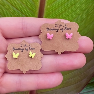 May include: Two pairs of butterfly-shaped stud earrings displayed on brown scalloped cardboard cards. One pair is bright yellow and the other is light pink. Each card features the text Thinking of Ewe with a small sheep icon.