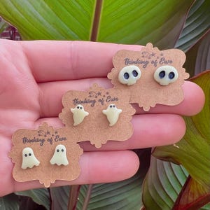 May include: Three pairs of Halloween-themed stud earrings. One pair features skull designs with dark blue eyes, and the other two pairs have ghost shapes. Each pair is displayed on a brown card with the text "Thinking of Ewe".