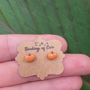 May include: A pair of orange pumpkin stud earrings on a brown card with the text "Thinking of Ewe". The earrings are small and detailed, ideal for autumn or Halloween. The card is held by a person.