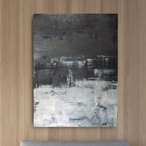 Op de afbeelding: Een grijs gestoffeerd bed met witte beddengoed en twee witte kussens. Het bed staat voor een houten muur met een groot abstract schilderij in zwart en wit.
