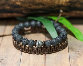 May include: Two bracelets are displayed. The top bracelet features dark gray lava rock beads. The bottom bracelet is a brown woven design. A silver and gray bead is centered on the top bracelet. The bracelets are on a wooden surface.