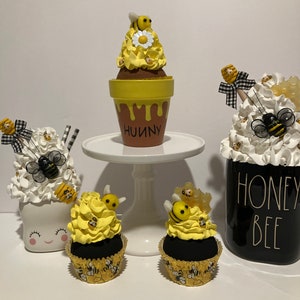 May include: A collection of decorative bee-themed items. Includes cupcakes with yellow frosting and bee toppers, a flower pot with the word "HUNNY", and a jar with "HONEY BEE" text. The items are decorated with bees and bows.