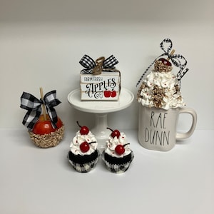 May include: A white Rae Dunn mug with black lettering that says "Rae Dunn" and a white cupcake with black and white checkered paper and a red cherry on top. There is a white cupcake with black and white checkered paper and a red cherry on top. There is a white cake stand with a wooden box that says "Farm Fresh Apples" with a red apple and a black and white checkered bow. There is a red apple with a black and white checkered bow and a brown and white topping.