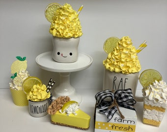 May include: A lemon-themed home decor display with a white mug with a yellow whipped cream topping and a lemon slice, a white mug with a yellow whipped cream topping and a lemon slice, a yellow and white pie, a yellow and white cupcake, a yellow and white cupcake, and a wooden block with the text "farm fresh lemons".