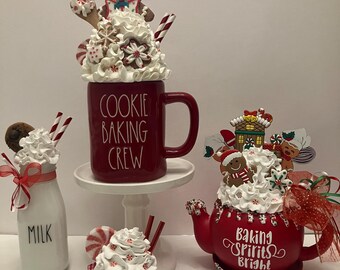 May include: A festive holiday display featuring a red mug with "COOKIE BAKING CREW" text, topped with whipped cream and gingerbread man. A red teapot with "Baking Spirits Bright" text, a milk bottle, and a small mug are also decorated.