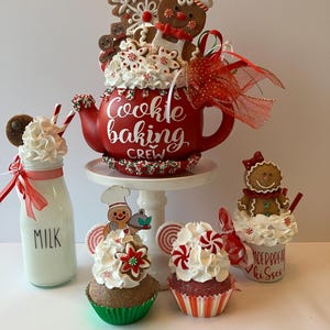 May include: A red teapot labeled Cookie baking CREW topped with gingerbread figures and faux whipped cream, accompanied by a milk bottle, two cupcakes with peppermint accents, and a small mug labeled GINGERBREAD kisses, all featuring festive holiday decorations.