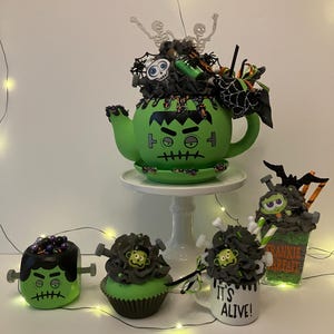 May include: A green teapot painted like Frankenstein's monster, topped with black decorations, skeletons, and a skull. Beside it are a matching green mug, a cupcake, a mug with the text "IT'S ALIVE!", and a glass with "FRANKIE PARFAIT" text.