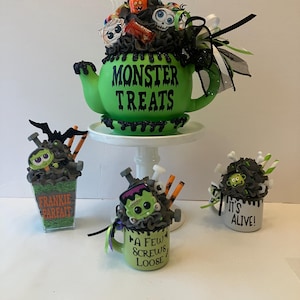 May include: A lime green teapot labeled MONSTER TREATS filled with Halloween decorations, alongside a square cup labeled FRANKIE PARFAIT, a mug labeled A FEW SCREWS LOOSE, and a mug labeled IT'S ALIVE, all featuring dark gray ruffles and monster accents.