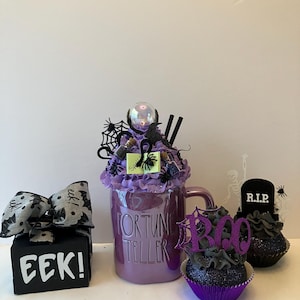 May include: A Halloween-themed display featuring a purple mug with "FORTUNE TELLER" text, a small black box with "EEK!" text, and cupcakes. The mug is decorated with a crystal ball, spiders, and a crow. The cupcakes have "BOO" and "R.I.P." toppers.