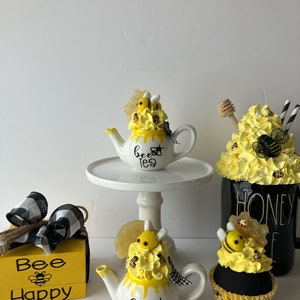 May include: Two white teapots with yellow and black bee decorations, a yellow and black cupcake, and a yellow wooden box with the text "Bee Happy" and a bee graphic. The teapots have the text "bee tea" on them.