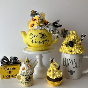 May include: A yellow teapot with the text "Bee Happy" and a bee design, decorated with yellow flowers, bees, and honeycomb.  A yellow mug with the text "Bee Happy" and a bee design, a yellow cupcake with bees, and a white mug with the text "Bumble" and a bee design.