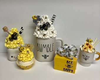 May include: A set of bee-themed home decor items, including a white ceramic mug with the word "Bumble" and a bee design, a yellow cupcake with a bee design, a small wooden box with the words "Bee Happy" and a bee design, and a white ceramic mug with a bee design and the words "Bee Happy".