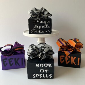May include: Three decorative wooden blocks with Halloween themes. One black block on a white stand reads Magic Spells Potions. A purple block reads EEK! and a black block reads BOOK OF SPELLS. Each block is topped with a decorative fabric bow.