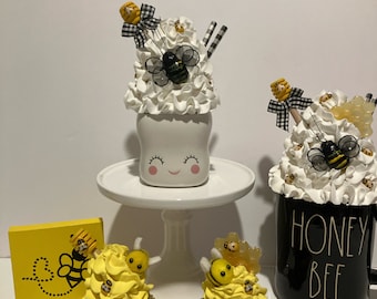 May include: Two yellow and black bee-themed cupcakes with white frosting and bee decorations. A white mug with a smiling face and white frosting with bee decorations. A black mug with white frosting and bee decorations. The black mug has the text "Honey Bee" on it. A yellow sign with a bee and honeycomb design.